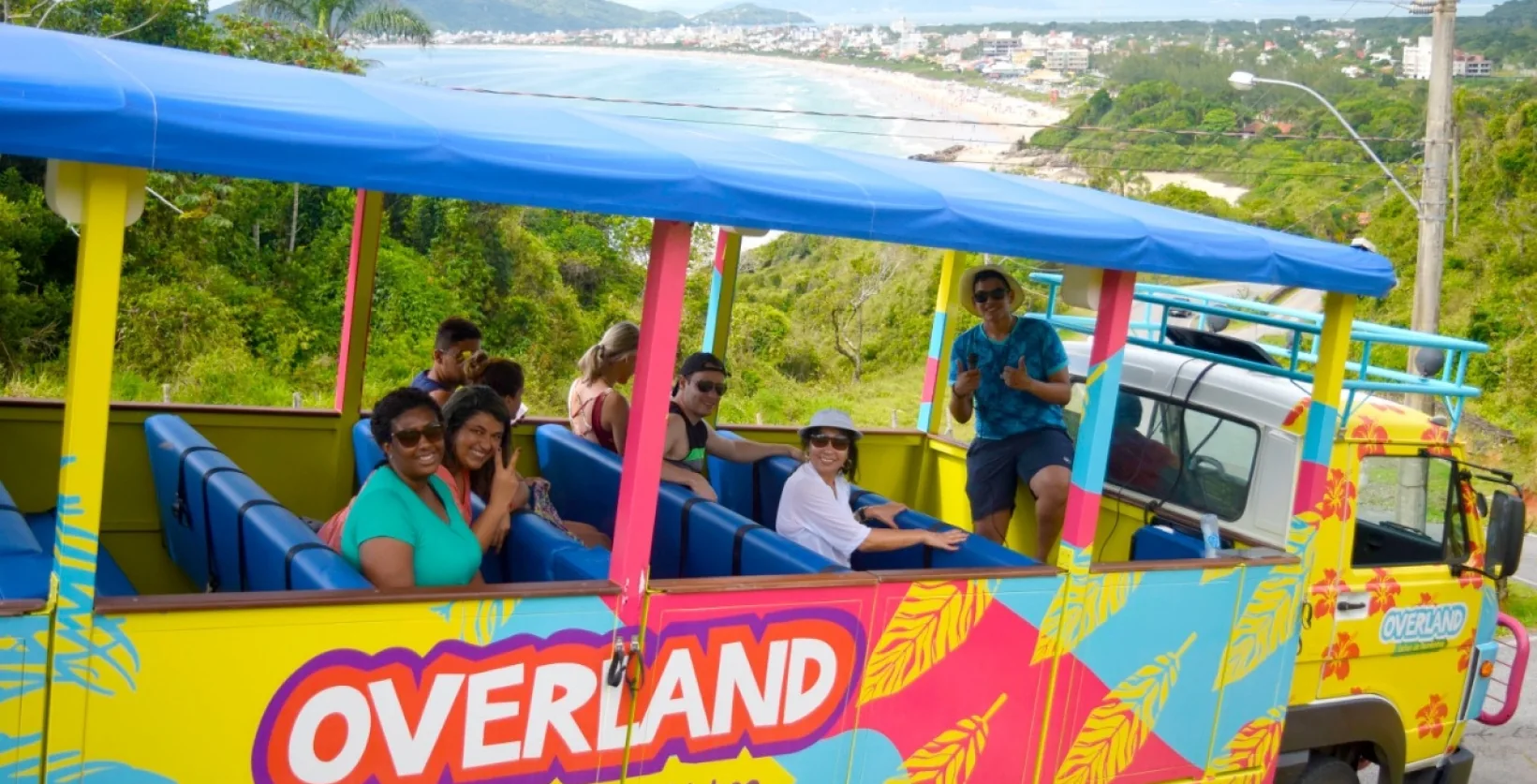 Turistas sorrindo a bordo de um veículo colorido do City Tour Safari em Bombinhas, com vista panorâmica para as praias e vegetação da região.