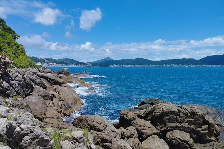 Vista da Trilha da Sepultura em Bombinhas, mostrando formações rochosas ao longo da costa, águas cristalinas e um horizonte montanhoso ao fundo, ideal para contemplar a natureza.
