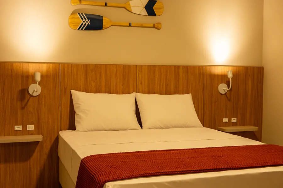 Cama de casal no quarto da pousada, com roupa de cama branca, cabeceira de madeira e iluminação aconchegante. Um espaço ideal para uma estada confortável e relaxante.