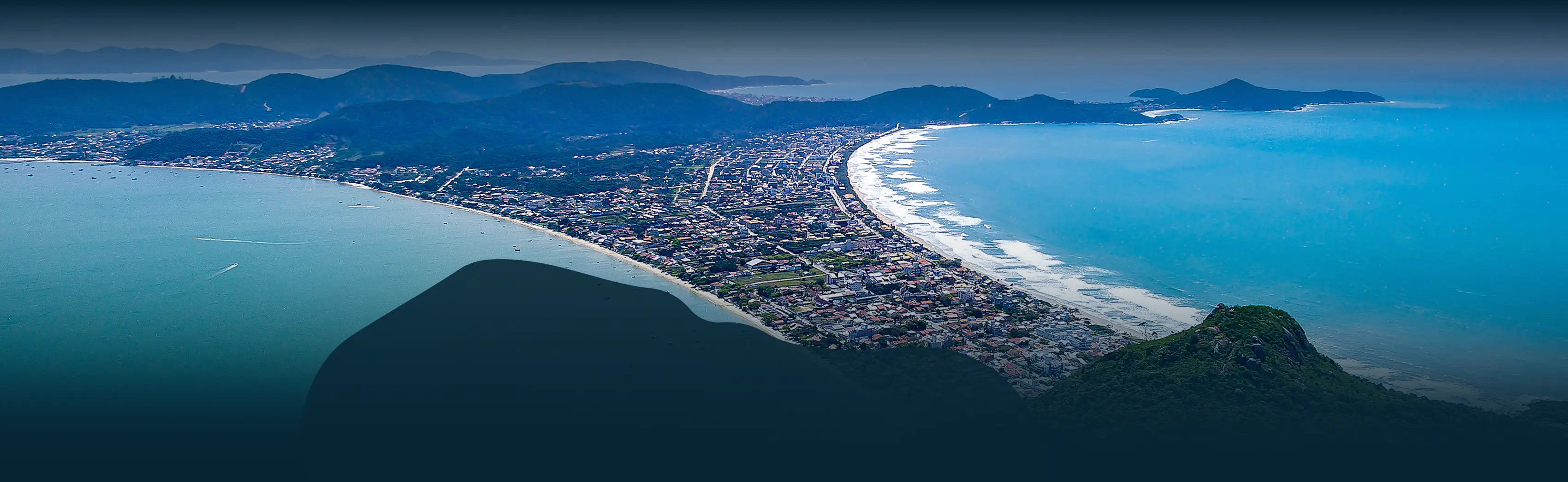 Vista aérea de Bombinhas, Santa Catarina, mostrando a praia de Canto Grande e a região montanhosa ao redor, com águas azuis e vegetação exuberante ao fundo.