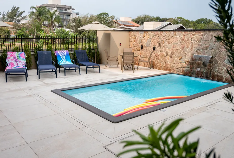 Área externa da Pousada Caminho do Bosque, com piscina rodeada por espreguiçadeiras, mesa com cadeiras e uma charmosa cascata, perfeita para momentos de lazer e relaxamento ao ar livre.
